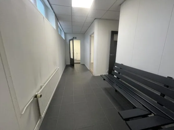 Gang met zwarte tegelvloer, witte muren, een zwarte bank en openstaande deuren in een gebouw aan de Burgemeester Smitsstraat.